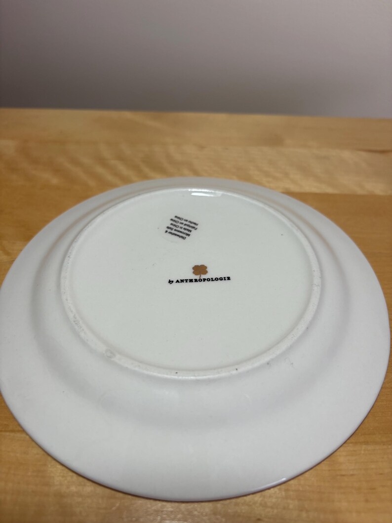 May include: White ceramic plate with a slightly raised rim. The underside features the text "by ANTHROPOLOGIE" in black, along with a small floral design and a sticker. The plate is on a light brown wooden surface.
