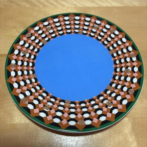 May include: A decorative plate with a blue center and a patterned border. The border features a geometric design with black, white, brown, and pink shapes, outlined in green. The plate is set on a wooden surface.