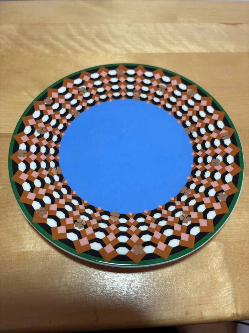 May include: A decorative plate with a blue center and a patterned border. The border features a geometric design with black, white, brown, and pink shapes, outlined in green. The plate is set on a wooden surface.