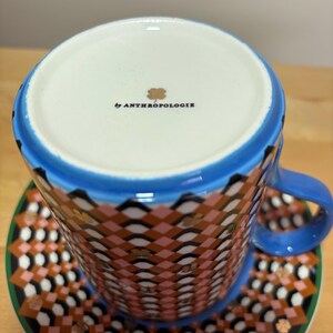 May include: A patterned teacup and saucer set. The cup and saucer feature a geometric design in shades of pink, brown, black, and gold. The cup has a blue rim and handle. The saucer has a green border. The bottom of the cup reads "by ANTHROPOLOGIE".