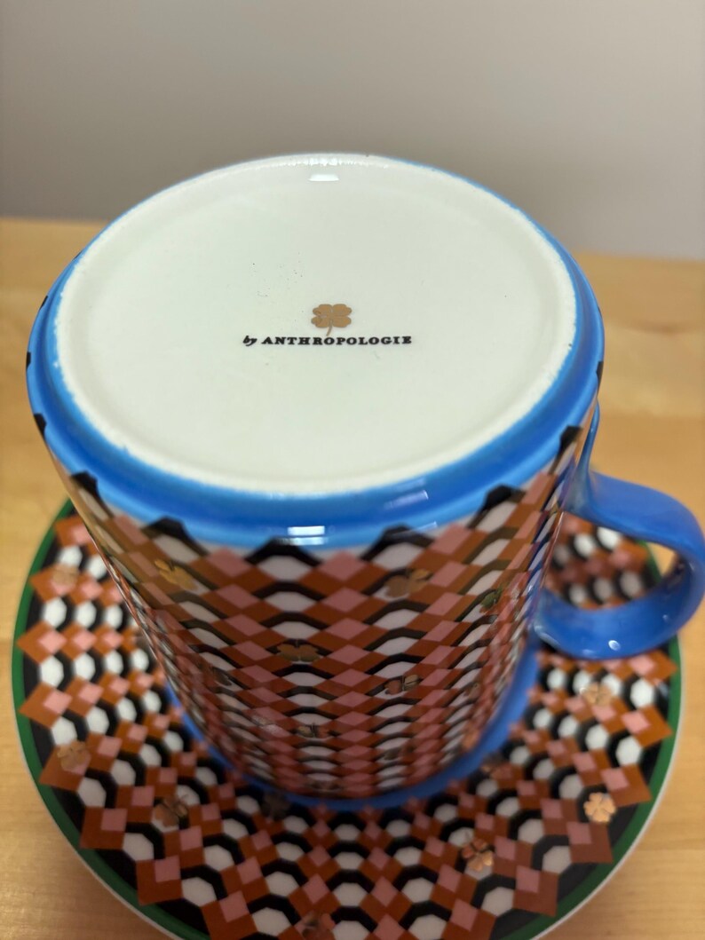 May include: A patterned teacup and saucer set. The cup and saucer feature a geometric design in shades of pink, brown, black, and gold. The cup has a blue rim and handle. The saucer has a green border. The bottom of the cup reads "by ANTHROPOLOGIE".