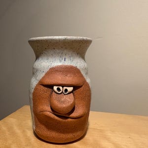 May include: A handmade ceramic vase with a face design. The vase features a light gray speckled top and a reddish-brown face with a nose, eyes, and a smiling mouth. The vase is a unique decorative item.