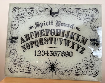 Könnte beinhalten: Ein rechteckiges Spirit-Board mit hellgrünem Hintergrund und schwarzen Druckgrafiken von drei Totenköpfen, einer Spinne und komplexen Spinnennetzen. Das Board zeigt das Alphabet in zwei Reihen, die Zahlen 0 bis 9 und den Schriftzug Spirit Board.