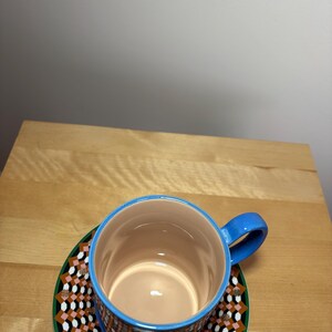 May include: A ceramic coffee cup with a blue handle and rim, resting on a matching saucer. The cup and saucer feature a geometric pattern in shades of brown, pink, black, and white, set against a green border. The interior of the cup is a light tan color.