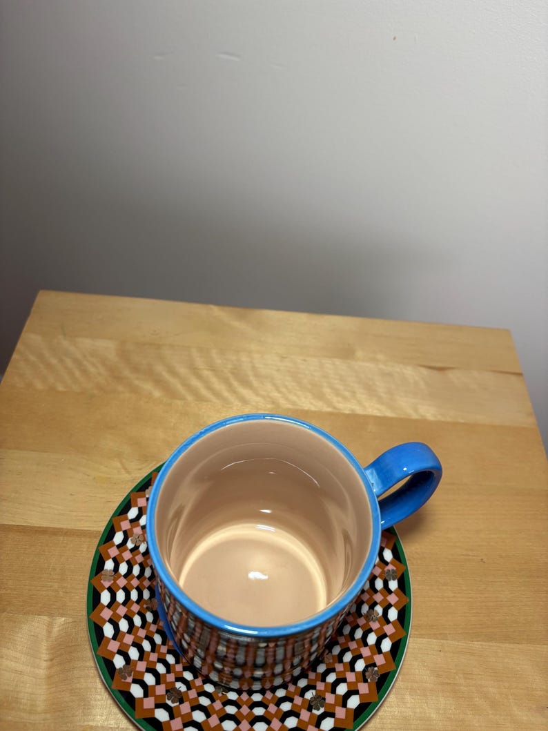 May include: A ceramic coffee cup with a blue handle and rim, resting on a matching saucer. The cup and saucer feature a geometric pattern in shades of brown, pink, black, and white, set against a green border. The interior of the cup is a light tan color.