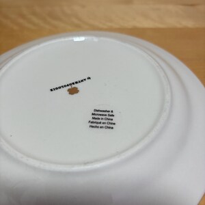 May include: A white ceramic plate with a slightly curved rim. The underside features the text "by ANTHROPOLOGIE" and a small floral design. Additional text indicates the plate is dishwasher and microwave safe, made in China.