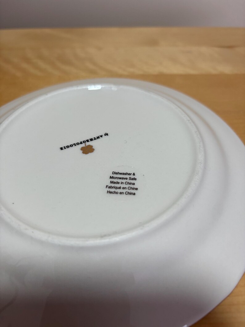 May include: A white ceramic plate with a slightly curved rim. The underside features the text "by ANTHROPOLOGIE" and a small floral design. Additional text indicates the plate is dishwasher and microwave safe, made in China.