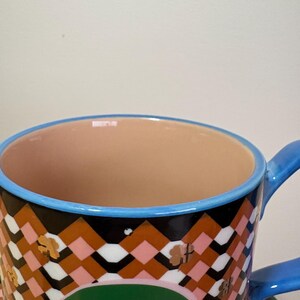 May include: A close-up of a ceramic cup with a blue handle and rim. The cup features a geometric pattern in brown, black, pink, and white, with gold clover accents. The interior is a light peach color.