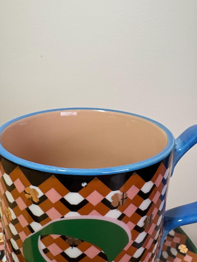 May include: A close-up of a ceramic cup with a blue handle and rim. The cup features a geometric pattern in brown, black, pink, and white, with gold clover accents. The interior is a light peach color.