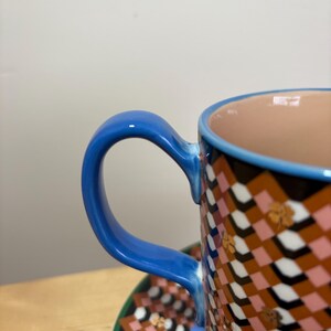 May include: Close-up of a patterned ceramic teacup and saucer. The cup has a blue handle and rim, with a geometric pattern in brown, pink, and white. The saucer mirrors the pattern with a green border.