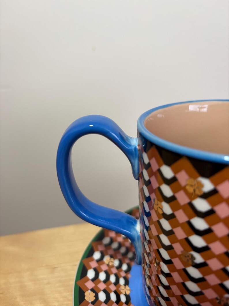 May include: Close-up of a patterned ceramic teacup and saucer. The cup has a blue handle and rim, with a geometric pattern in brown, pink, and white. The saucer mirrors the pattern with a green border.