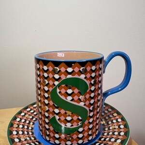 May include: A patterned ceramic mug with a large green "S" and a matching saucer. The mug has a blue handle and a pink, black, and white geometric design. The saucer has a similar pattern with a green border.