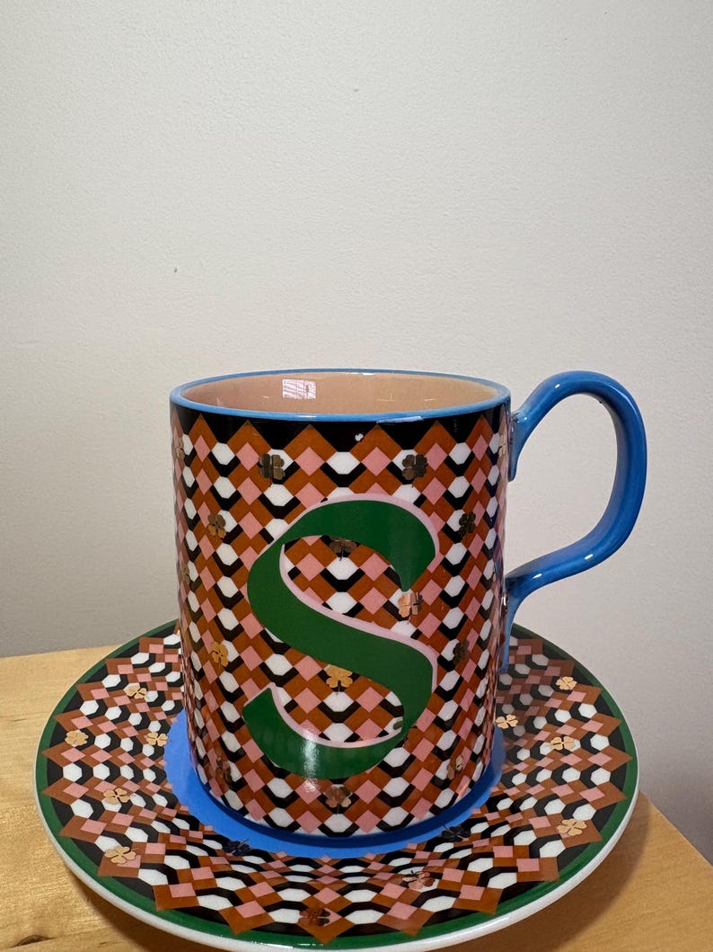 May include: A patterned ceramic mug with a large green "S" and a matching saucer. The mug has a blue handle and a pink, black, and white geometric design. The saucer has a similar pattern with a green border.