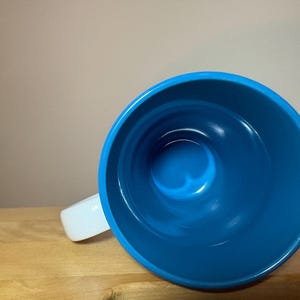 以下が含まれることがあります： 鮮やかな青い内側と白い外側を持つセラミック製のマグカップ。真上から見た状態で、円形の開口部、青い内壁の滑らかで光沢のある仕上げ、そして左側に伸びる白いハンドルが確認できます。