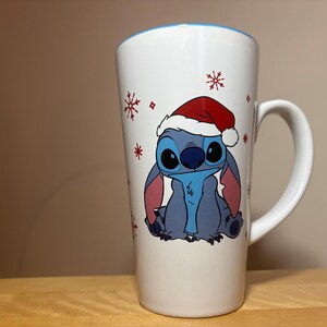 以下が含まれることがあります： 赤いサンタ帽子をかぶった青いキャラクター、スティッチのグラフィックが描かれた背の高い白いセラミックマグカップ。デザインには、キャラクターの周りに小さな赤い雪の結晶の模様が散りばめられています。マグカップの内側の縁は水色です。