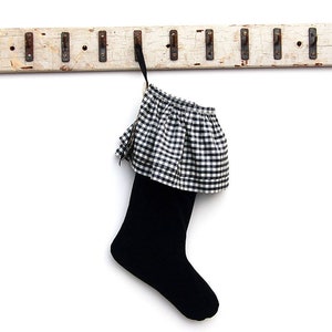 May include: A black Christmas stocking with a black and white checked frill, suspended from a distressed, white wooden rack with metal hooks. The rack is mounted on a white wall.
