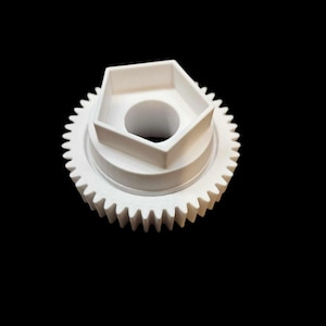 Peut inclure: Une pièce d'engrenage blanche imprimée en 3D, avec un dessus hexagonal et un trou central. L'engrenage a un bord extérieur denté et est posé sur un fond noir. L'objet semble être un composant mécanique.