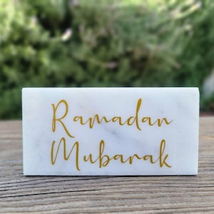 May include: A white marble block with the gold text "Ramadan Mubarak". The marble has grey veining. The background is a blurred view of green foliage. The block is rectangular in shape.