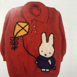 Könnte beinhalten: Roter Strickpullover mit einem Cartoon-Hasen, der einen Drachen hält. Der Drachen ist gelb mit schwarzer Umrandung, und der Hase trägt ein blaues Outfit. Der Pullover hat einen Kragen und zwei Knöpfe.