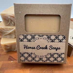 May include: A stack of handmade goat's milk soaps with a kraft paper box. The box has a window showing the soap and a label that reads "Horse Creek Soaps". The soaps are light brown and appear to be unscented.