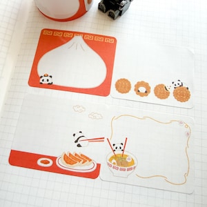May include: Four white notepads with red borders and food-themed illustrations. One notepad features a dumpling with a panda, another has mooncakes and a panda, and the other two have dumplings and ramen with pandas. The notepads are on a grid-lined paper.