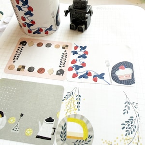 May include: A collection of illustrated stationery items featuring a strawberry and leaf design. Includes a ceramic mug, rectangular cards with food and house illustrations, and a small robot figurine with the text "YOU LOVE". The color palette is soft and pastel.