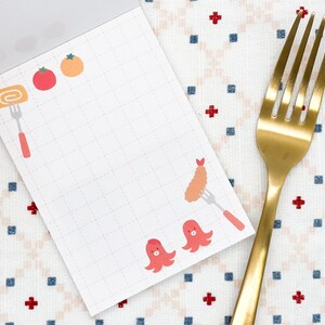 May include: A notepad with a grid pattern and food illustrations, including a roll, tomatoes, oranges, tempura, and octopus. A gold fork rests beside the notepad on a patterned fabric.