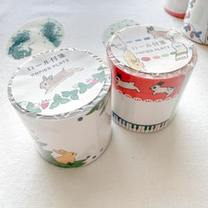 May include: Two rolls of decorative tape with bunny and strawberry designs. One roll features a green and white pattern with a bunny and strawberries. The other roll has a red and white design with bunnies and a piano key border. The text "PAPIER PLATZ" is visible.