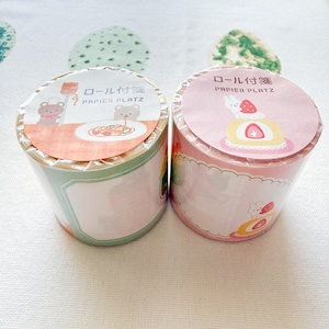 May include: Two rolls of decorative tape with Japanese text and cute illustrations. One roll features a bear chef with pasta, the other a bunny with a strawberry cake. The tape rolls are in shades of pink, green, and white.