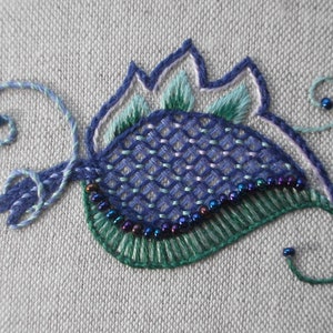May include: Embroidered floral design in shades of blue, green, and white on a light grey fabric. The design features a central flower shape with intricate stitching and beaded accents. The embroidery is detailed and textured.