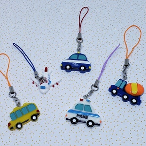 Op de afbeelding: Vijf kleurrijke sleutelhangers met miniatuurvoertuigen. De sleutelhangers omvatten een blauwe en witte politieauto, een blauwe en witte taxi, een wit vliegtuig, een blauwe en oranje cementwagen en een gele schoolbus.