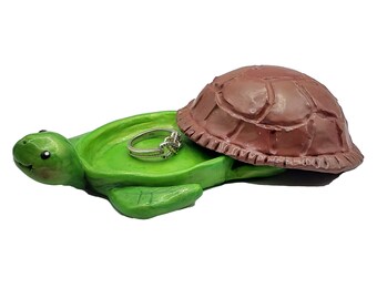 Puede incluir: Una caja de joyas con forma de tortuga verde con una tapa de caparazón marrón. La tapa está abierta y un anillo de plata está dentro de la caja.