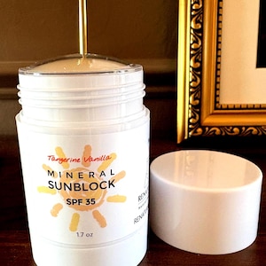 May include: White cylindrical mineral sunblock container with the text "Tangerine Vanilla MINERAL SUNBLOCK SPF 35" and a sun graphic. The container is 1.7 oz. The cap is off, revealing the product. A gold-colored metal rod is visible.
