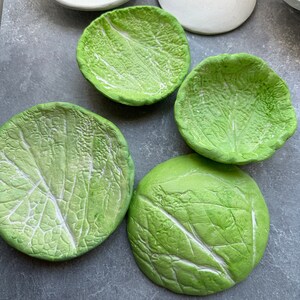 Puede incluir: Cuatro platos de cerámica verde con un diseño de hoja de col. Los platos son redondos y tienen un borde ligeramente elevado.
