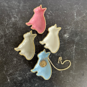 May include: Four ceramic cat-shaped dishes in pink, white, and blue. The blue dish has a gold chain and a gold coin pendant.