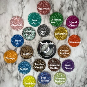 May include: A collection of 18 small round jars with different colored lids and labels. The jars are filled with a variety of scented wax melts. The labels include names like "Peppermint", "New Car", "Christmas Tree", "Island Citrus", "Vanilla Pound Cake", "Hot Apple Pie", "Amoretto", "Cinnamon", "Black Ice", "Leather", "Orange", "Cowboy Britches", "Leather Vanilla", "Ocean", "Caramel Vanilla", "Black Cherry", "Baby Powder", "Christmas Traditions", and "Jasmine".