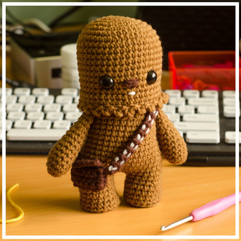 May include: A brown crocheted Chewbacca character with a white bandolier and a brown pouch. The character has black eyes and a white mouth.