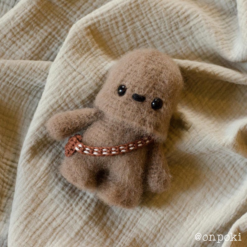 May include: A brown crocheted Chewbacca plush toy with black eyes and a brown and white braided belt. The toy is sitting on a light brown fabric background.