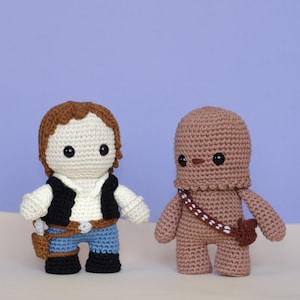 May include: Two crocheted plush toys, one brown and one white with brown hair, standing side by side on a light brown surface. The white toy is wearing a black vest, a white shirt, blue jeans, and a brown belt with a holster. The brown toy has a brown strap across its chest.