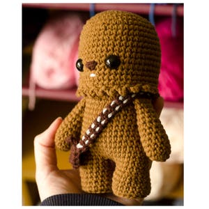 May include: A brown crocheted Chewbacca plush toy with black eyes and a white and brown bandolier. The toy is being held in a person's hand.