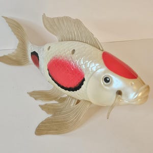 May include: A plastic koi fish toy with a pearlescent white body, red and black markings, and translucent fins. The fish has a detailed design, including fins, tail, and whiskers, and is likely a decorative item or toy.