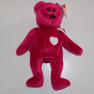May include: A pink Beanie Baby teddy bear with a white heart on its chest. The bear has black eyes and a black nose.