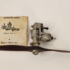 May include: An antique Rogers RMC 2 model airplane engine with a wooden propeller. The engine is silver and black, and the propeller is brown. An instruction manual is also present, with the text "Rogers RMC 2" and "Rogers Motor Co."