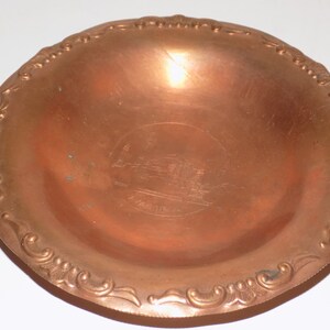 May include: A small, copper-colored trinket dish with an ornate, raised rim and a design of a building in the center. The dish is about 5 inches in diameter.
