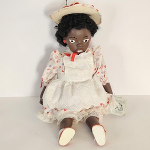 May include: A vintage doll with dark skin, wearing a white eyelet dress with red accents and a matching hat. The doll has black curly hair, red earrings, and red shoes. A tag is attached to the doll.