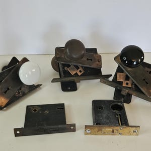 May include: An assortment of antique door hardware, including doorknobs and mortise locks. The doorknobs are white, brown, and black. The locks are made of metal with keyholes and mounting plates. The hardware is likely from an older home.