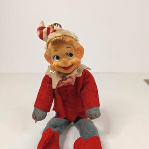 May include: A vintage elf doll with a molded plastic face, blue eyes, and a smiling expression. The doll wears a red felt jacket, gray felt pants, red felt boots, and a white collar with gold glitter accents. A red and white striped hat sits atop.