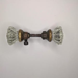 May include: A vintage-style door knob set featuring two clear, fluted glass knobs connected by a central metal spindle. The hardware includes aged brass-toned circular bases and a visible set screw on one side, providing a functional mechanism.