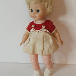 May include: A vintage doll with blonde hair, blue eyes, and a red and white dress. The doll is wearing white shoes and has a sweet expression on its face.