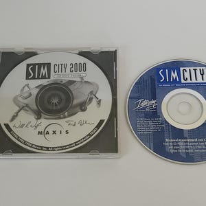 May include: Two computer game CDs. One CD is in a clear case with "SIM CITY 2000" printed on it, along with signatures and the Maxis logo. The other CD is blue with "SIM CITY" and the Interplay logo.